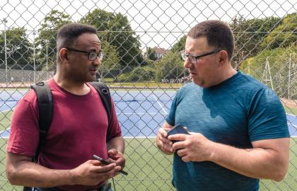 Two people facing each other talking, one is showing the other something on their phone. They are stood in front of the tennis courts at Hove Park.