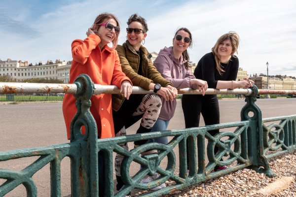 Four people stood in a row whilst leaning on railings and talking on a windy day on Hove seafront