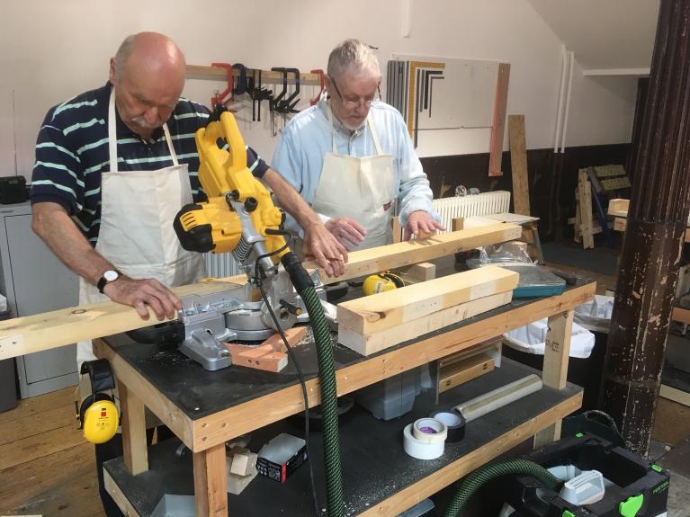 Photo of two men wearing aprons working on a woodwork project