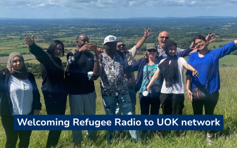 Photo of a group of people standing in a grassy field on a hilltop, arms raised in celebration, with rolling Sussex countryside visible in the background under…Photo of a group of people standing in a grassy field on a hilltop, arms raised in celebration, with rolling Sussex countryside visible in the background under a blue sky. Text overlay reads "Welcoming Refugee Radio to UOK network".