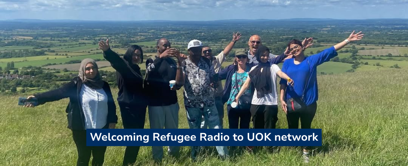 Photo of a group of people standing in a grassy field on a hilltop, arms raised in celebration, with rolling Sussex countryside visible in the background under…Photo of a group of people standing in a grassy field on a hilltop, arms raised in celebration, with rolling Sussex countryside visible in the background under a blue sky. Text overlay reads "Welcoming Refugee Radio to UOK network".