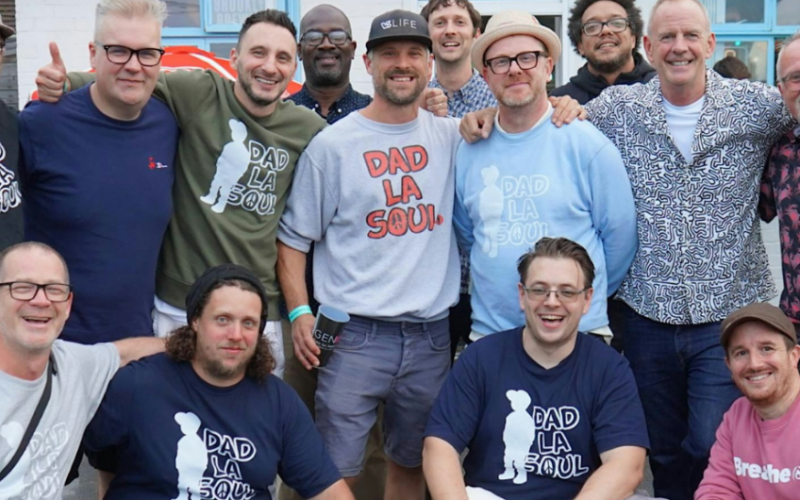 A group of fourteen smiling men, many wearing Dad La Soul T-shirts, stand and crouch together outside a building.