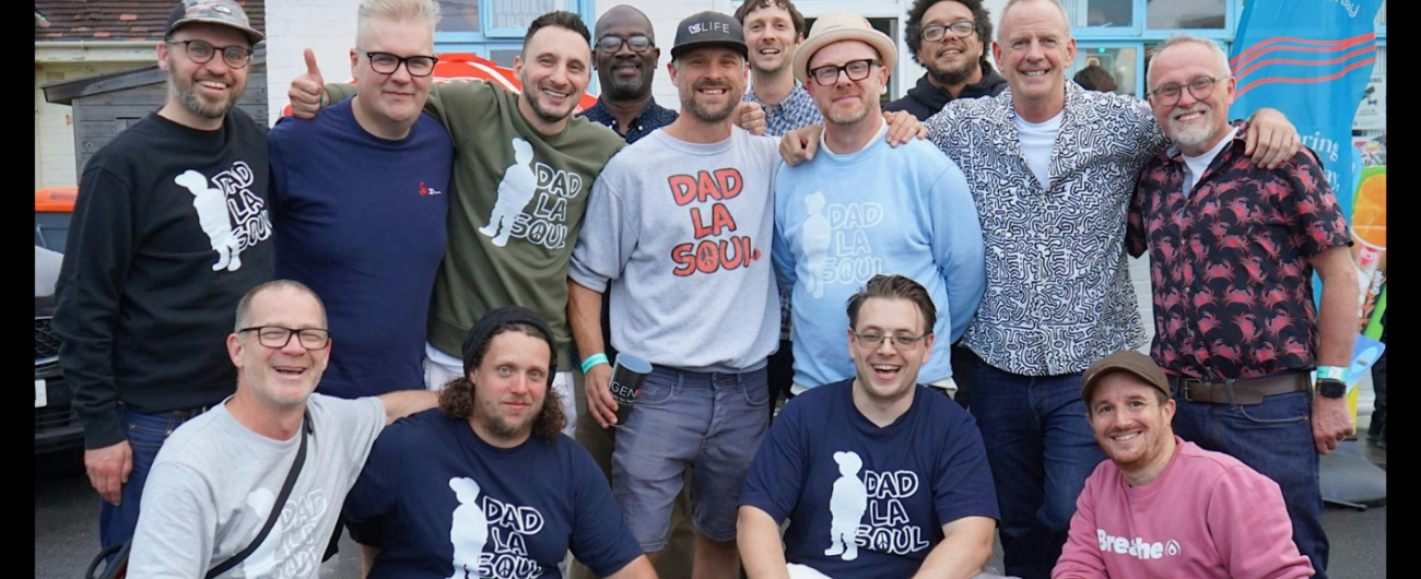 A group of fourteen smiling men, many wearing Dad La Soul T-shirts, stand and crouch together outside a building.