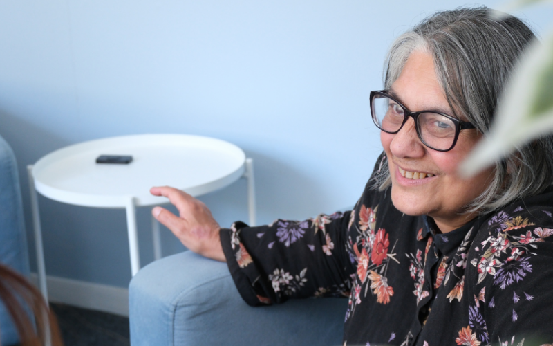 A woman sitting in a chair smiling, wearing glasses and a black floral shirt.