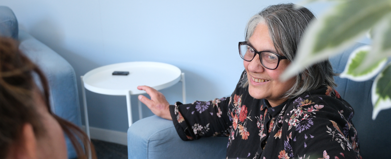 A woman sitting in a chair smiling, wearing glasses and a black floral shirt.